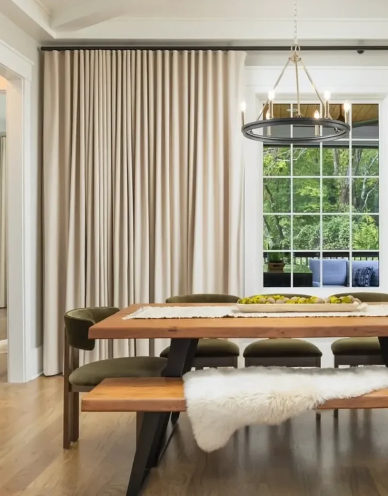 Dining room with a wooden table, green chairs, and a beige curtain. A modern chandelier hangs above; a large window shows a view of green trees outside.